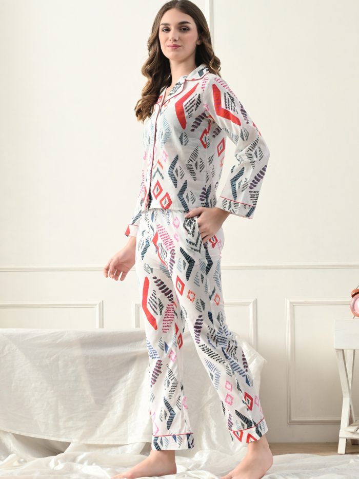 A woman standing in a bedroom wearing a long-sleeved white nightgown with a multicolor geometric print.